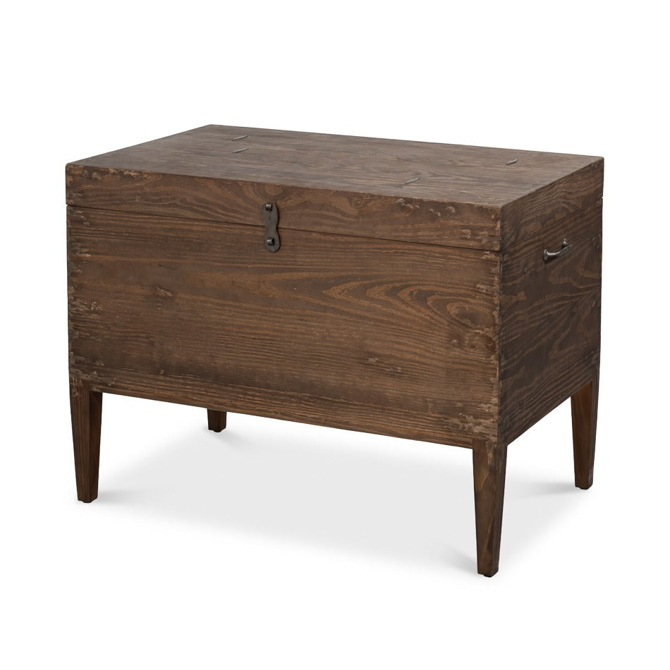 rustic trunk-form side table in antique creamy beige pine with a central iron clasp, side metal handles, and tapered dark wood legs