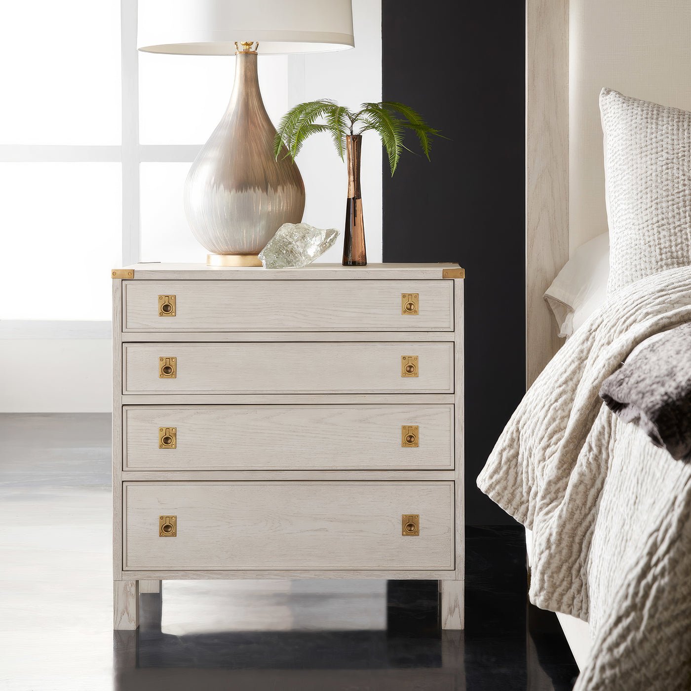 Campaign style nightstand beside bed with lamp and decor.
