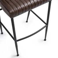 Close-up of the fluted brown leather bar stool’s stitched seat edge, black iron legs and curved footrest