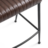 Close-up of the fluted brown leather counter stool’s stitched seat edge, black iron leg and curved footrest