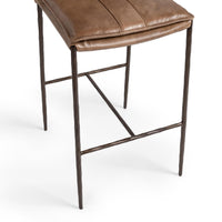 Close-up of the slim-frame tan leather bar stool’s padded seat edge, hammered-iron legs and footrest
