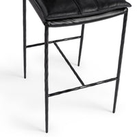 Close-up of the slim-frame black leather bar stool’s padded seat edge, hammered-iron legs and footrest
