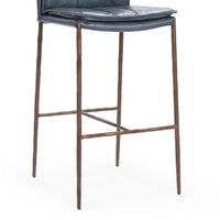 Close-up of the slim-frame blue leather bar stool’s padded seat edge, brown iron legs and footrest