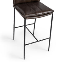 Close-up of the slim-frame dark brown leather bar stool’s padded seat edge, hammered-iron legs and footrest
