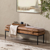 Tan leather bench with strap details styled beneath artwork with a fringed throw and a wooden tray