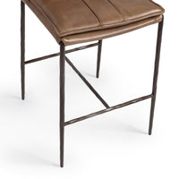 Close-up of the slim-frame tan leather counter stool’s padded seat edge, hammered-iron legs and footrest