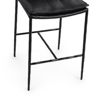 Close-up of the slim-frame black leather counter stool’s padded seat edge, hammered-iron legs and footrest