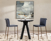 Two slim-frame blue leather counter stools around a round black table beneath abstract artwork