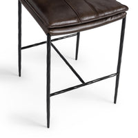Close-up of the slim-frame dark brown leather counter stool’s padded seat edge, hammered-iron legs and footrest